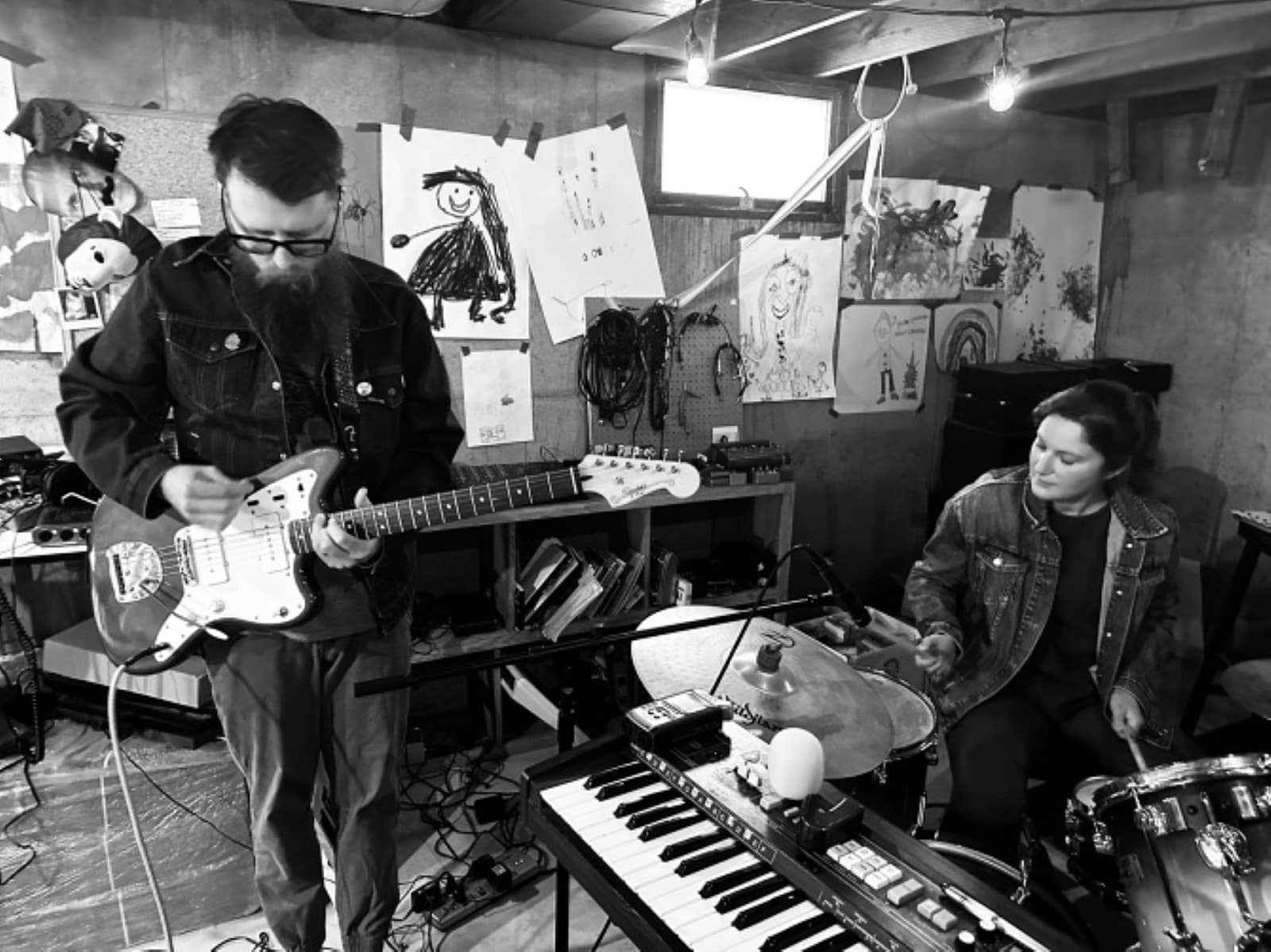 A black-and-white photo of rock duo Red Pants playing in a practice space. Jason Lambeth plays guitar while standing and looking down to the left of the image. Off to his right is drummer Elsa Nekola. A keyboard is visible at the bottom of the image, slanted towards Lambeth's waist. Both Red Pants members are in dark clothing. A series of Lambeth's visual art adorns the wall behind them.|