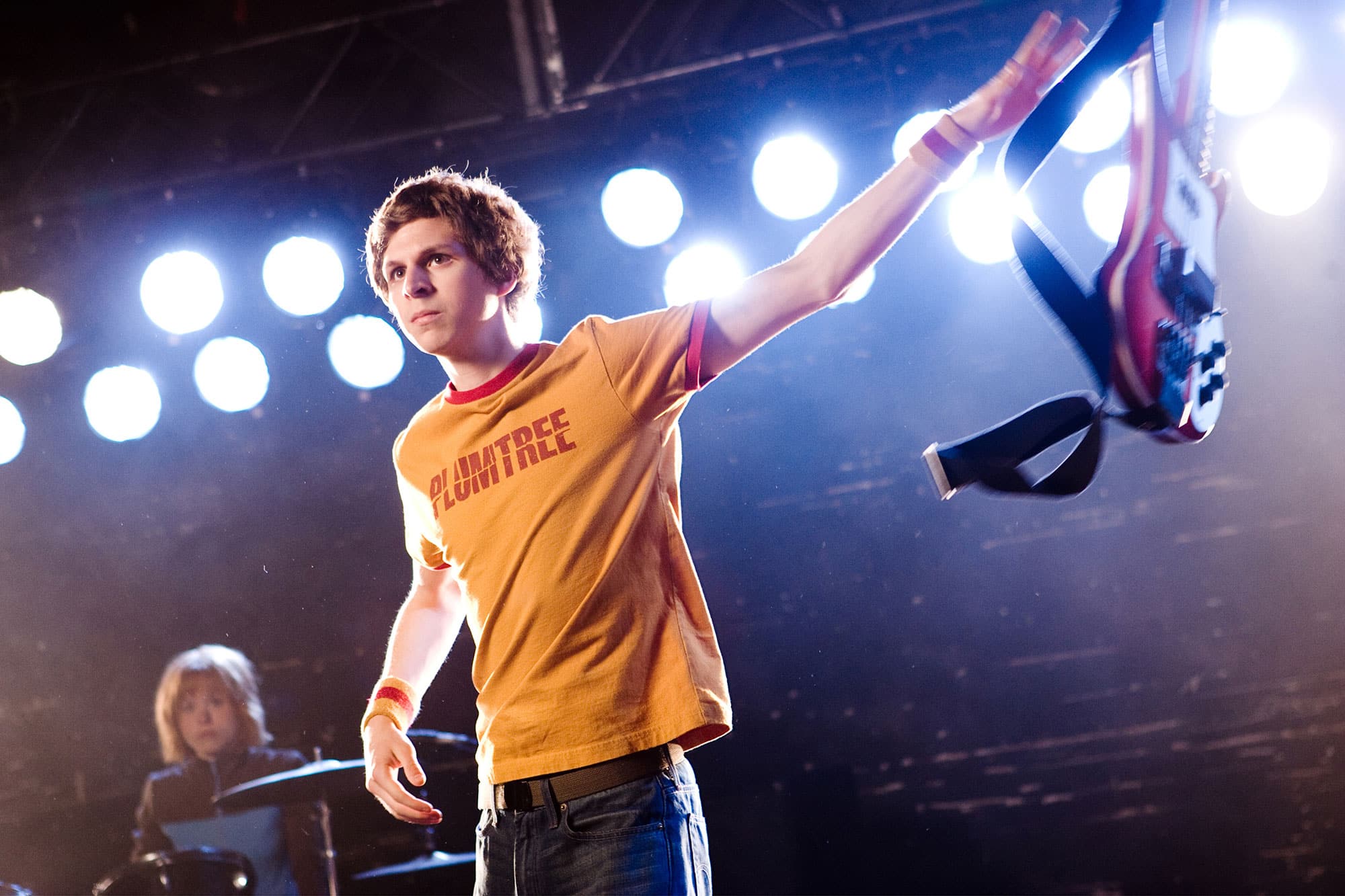 Scott Pilgrim throws a red Rickenbacker bass to the right side of the image. He's wearing a yellow-orange Plumtree t-shirt. Stage lights can be seen behind him, as well as his band's drummer.