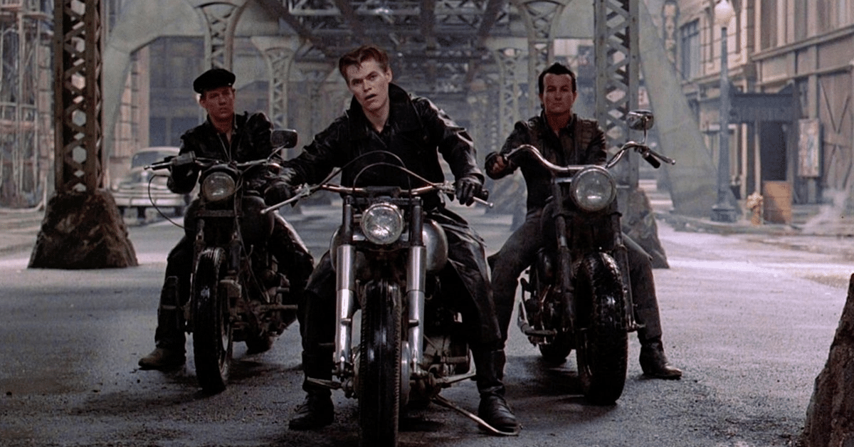 Three leather-clad men sit on motorcycles under a metal bridge at a medium shot. They look forward off-screen into the city streets in daylight.