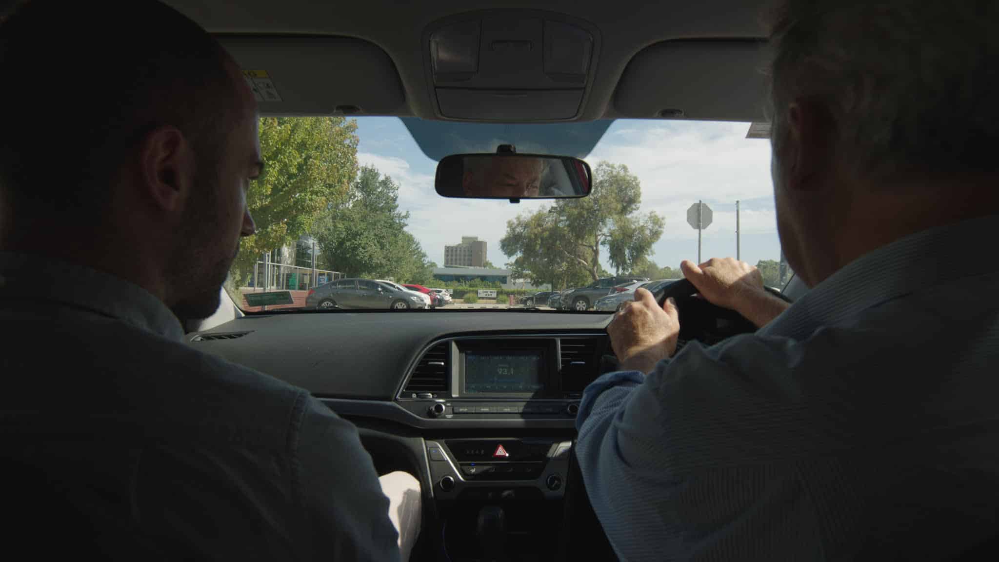 A shot from the backseat of Andrew Rakowski's Hyundai Elantra shows him (right) and director David Easteal in the passenger's seat (left) shortly after leaving their office park for the day.|A shot above the Rakowski's country estate from the perspective of a drone renders the landscape like the surface of a desert planet.|A production photo of the camera setup in the backseat of Andrew Rakowski's red Hyundai Elantra.||A headshot of director David Easteal.