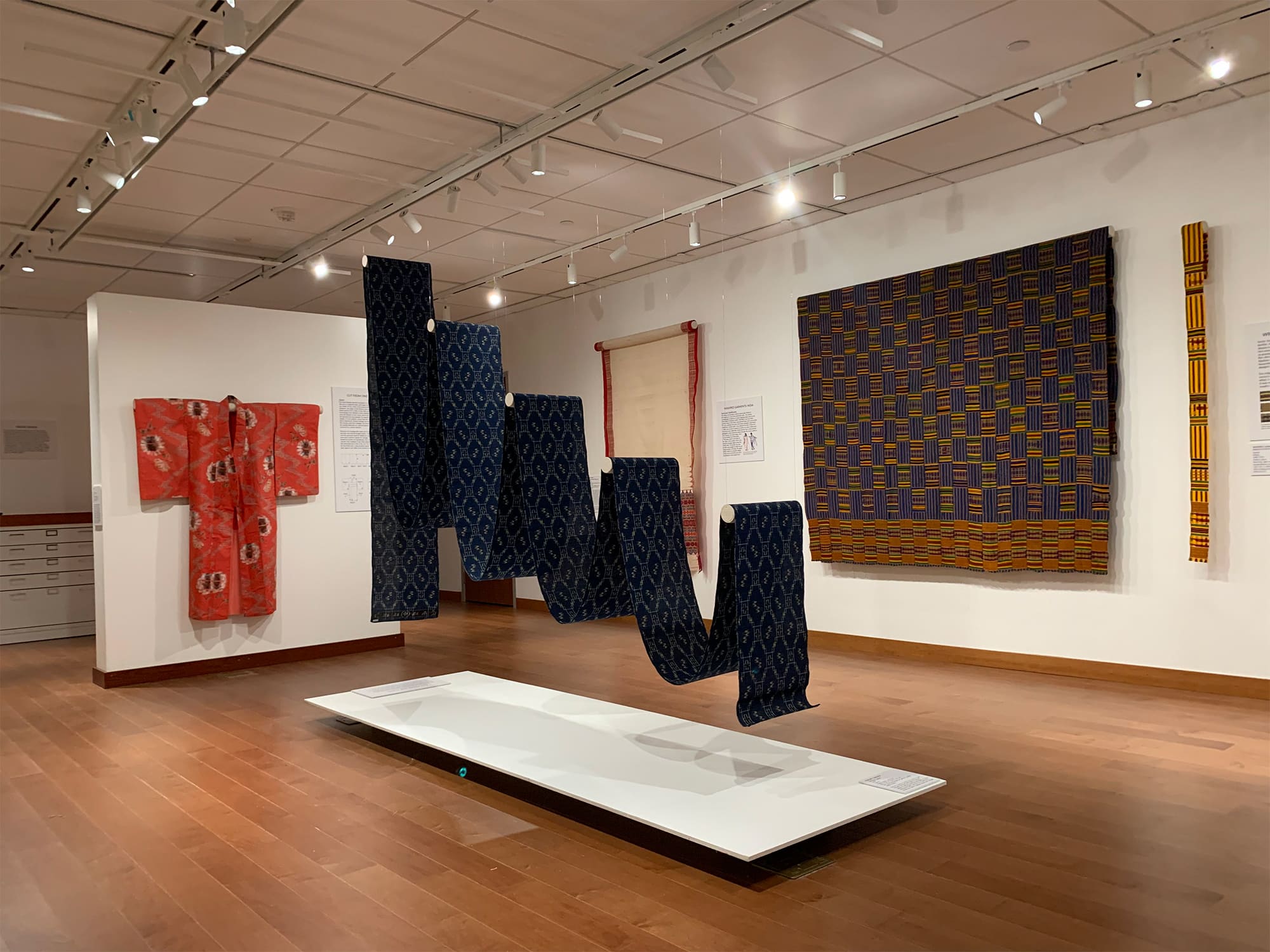 A photo of the Lynn Mecklenburg Textile Gallery shows several items in the "Uncut Attire" exhibit mounted on walls, and a long, blue piece suspended in cascading waves from the ceiling.|||