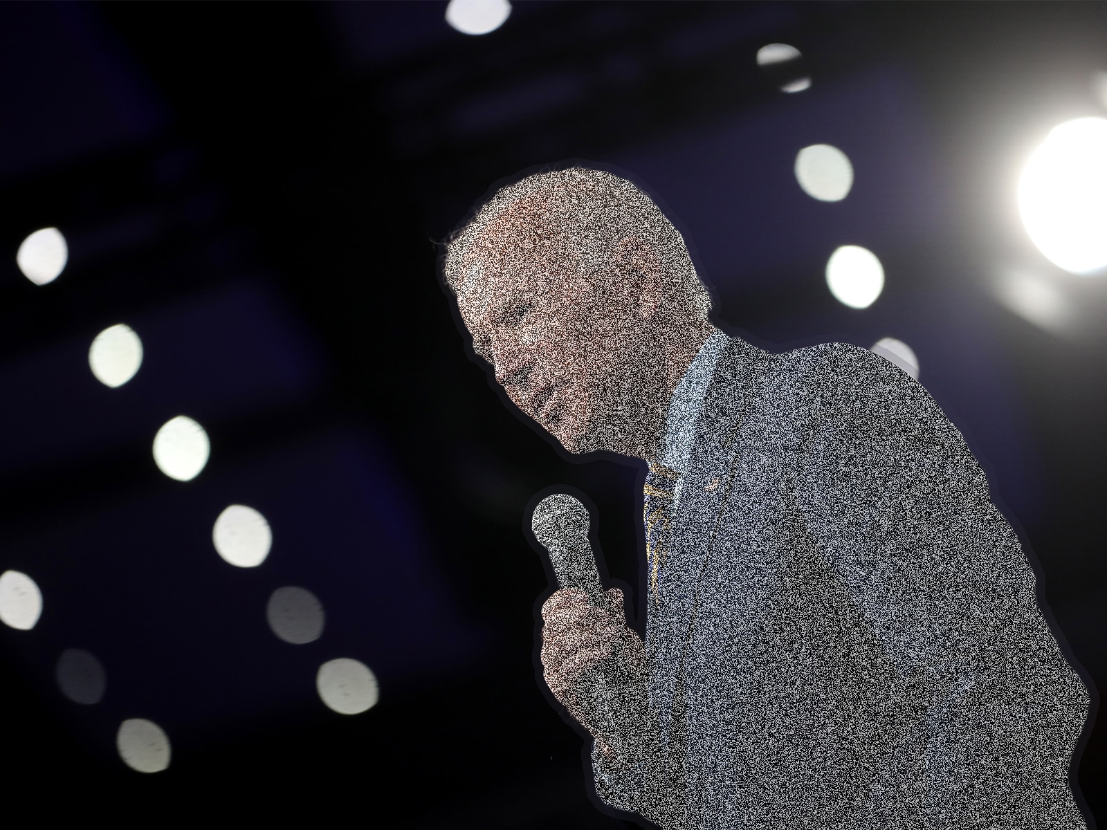 A photo shows Joe Biden speaking at a public event. Biden is shot from below, so that he looms over the camera at an angle, with bright stage lights behind him. The photo has been altered such that Biden is overlaid with a field of distorted static.