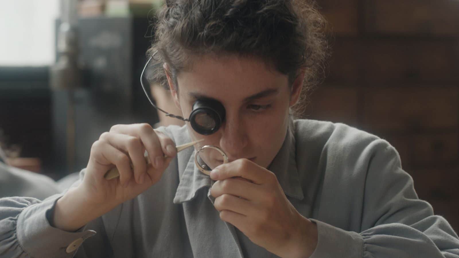 A young woman in a grey, long-sleeved dress shirt looks through a magnifying glass on her right eye at a watch mechanism.