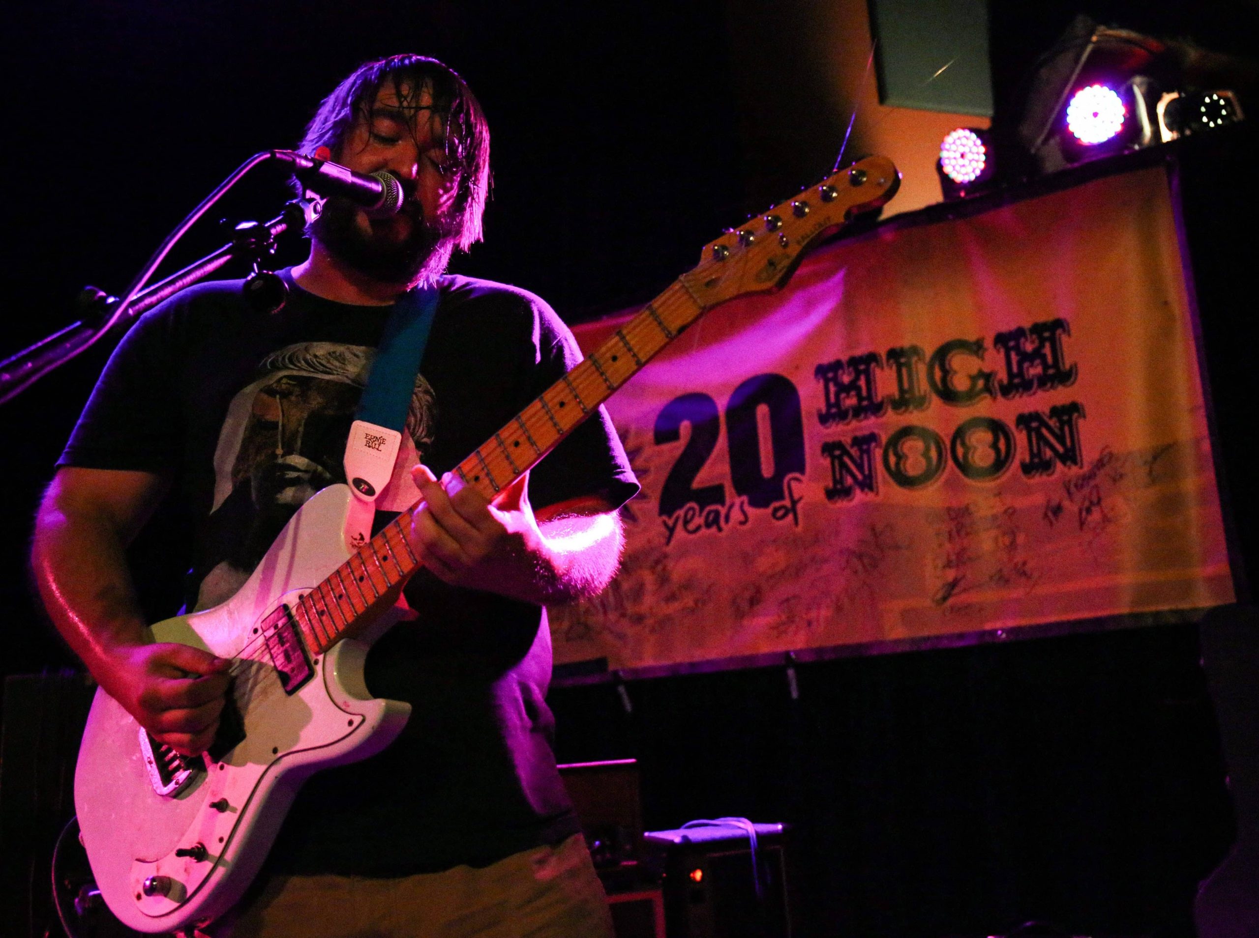 We Should Have Been DJs' Mike Pellino is pictured playing guitar and singing on stage at the High Noon Saloon in early June. He is to the left third of the image. The upper right segment displays a "20 Years of High Noon Saloon" banner adorning the wall of the venue, the banner bears the signatures of many bands. The image is dark and some film grain is evident, but the venue's stage lights provide a discernible blue/orange sheen.